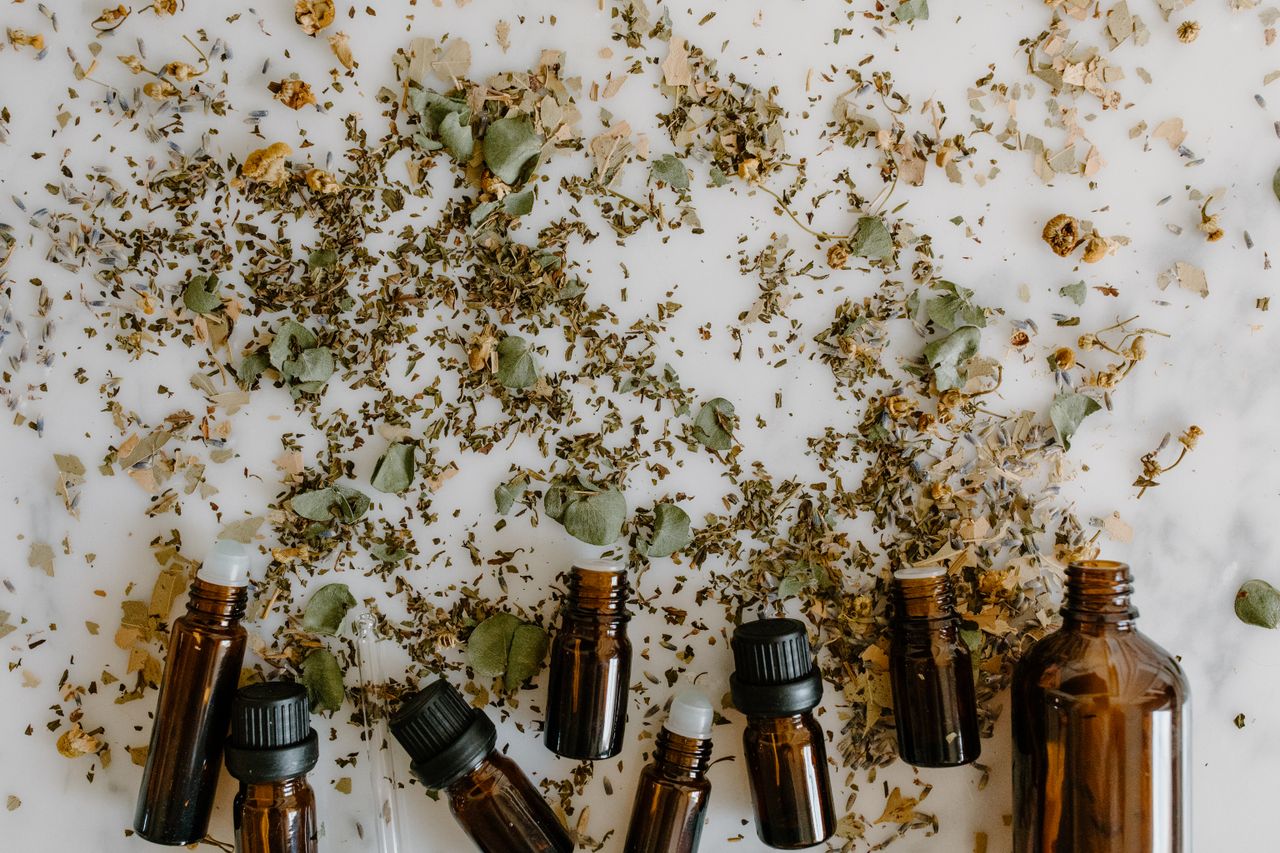Glass bottles with essential oils surrounded by scattered dried herbs on a white surface.